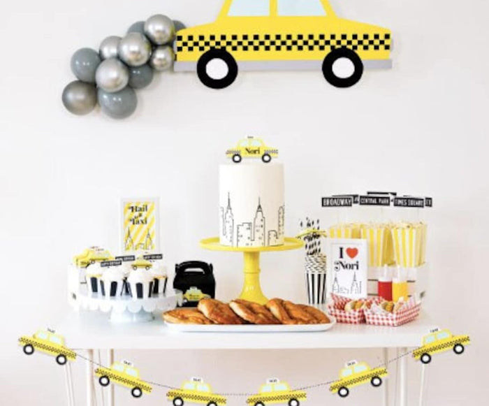 New York Taxi Wall Art Backdrop above a full dessert table with a silver balloon garland, a personalized skyline cake, pretzels, hot dogs, and a taxi garland