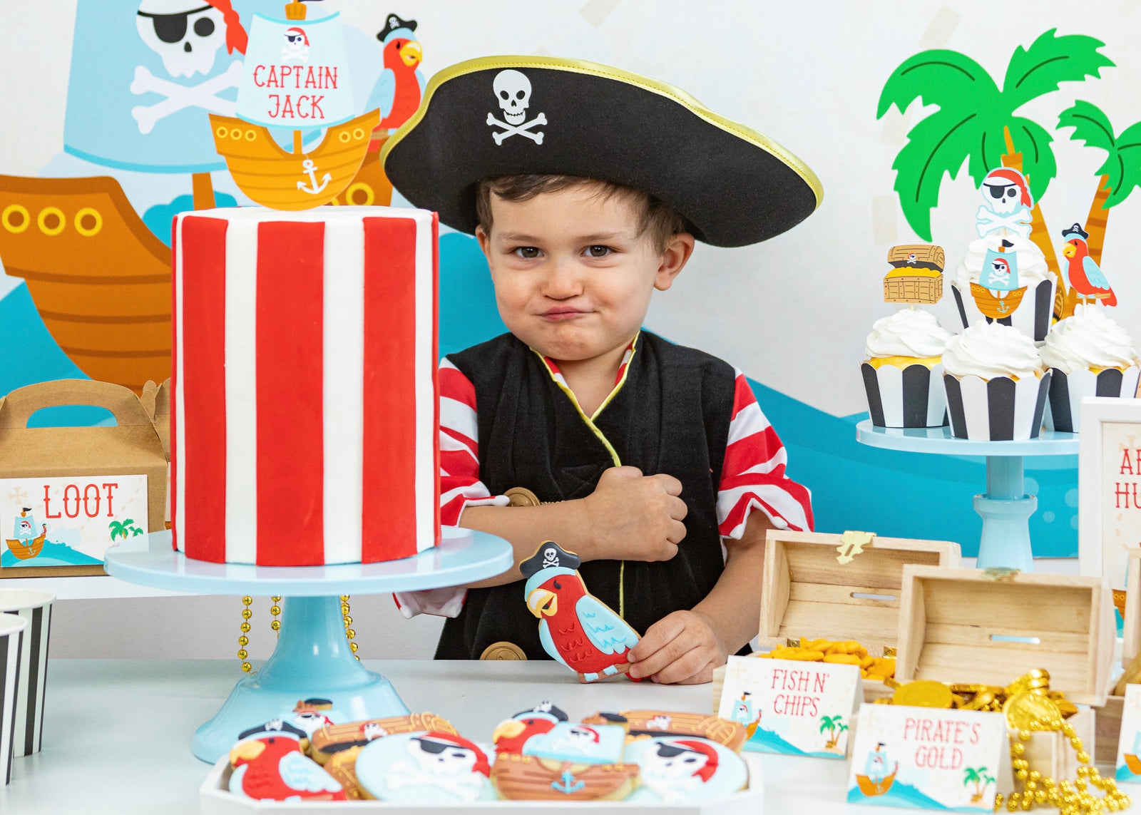 A child wearing a black pirate hat with a skull and crossbones sits behind a decorated pirate party table. The table displays a red and white striped cake on a blue pedestal, pirate cookies, small wooden treasure chests, and a cupcake stand with more pirate-themed cupcakes.