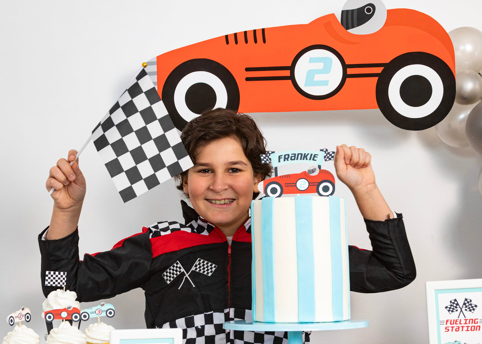 A child wearing a black and red racing jacket stands behind a decorated party table with a blue-and-white striped cake, car-shaped cupcake toppers, and race-themed party decorations including a large orange race car sign and checkered flags.