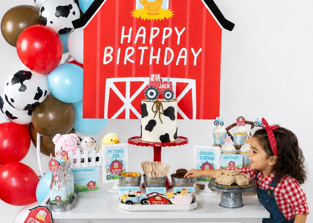 A children's farm-themed birthday party setup with a large red barn sign reading 'HAPPY BIRTHDAY,' a decorated table with cupcakes, balloons, and a banner, and a young girl in overalls standing beside the table.